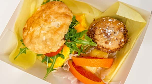 Breakfast box with a biscuit sandwich, scrambled eggs, arugula, muffin, and orange slices.