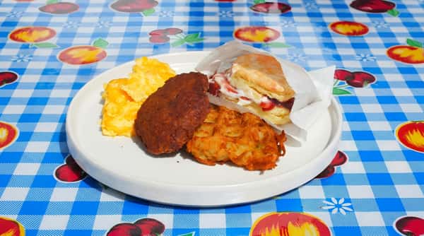 Breakfast plate with scrambled eggs, dark brown sausage patty, golden hash brown, and a biscuit with cream and jam in a paper sleeve.