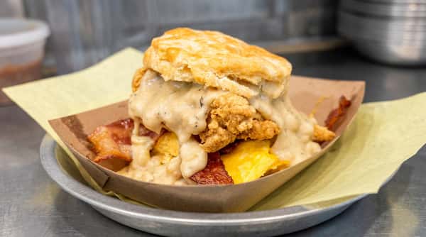 Biscuit sandwich with fried chicken, gravy, scrambled eggs, and bacon in a paper tray.