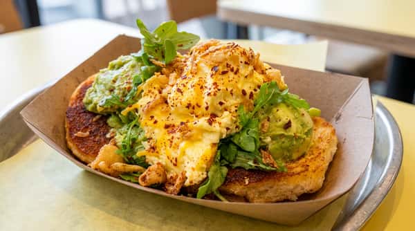Scrambled eggs with cheese, avocado, arugula, and chili flakes on two toasted bread slices in a cardboard tray.
