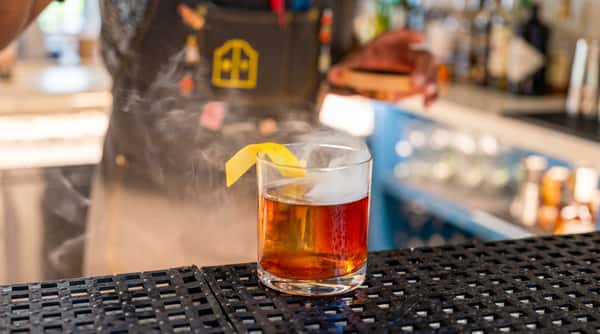 Cocktail in a short glass with amber liquid, lemon peel garnish, and smoke rising from the top.