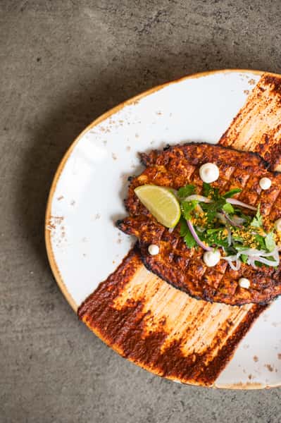 Grilled whole fish with chili rub, cilantro, red onion, lime, and white sauce dots on a rustic white plate