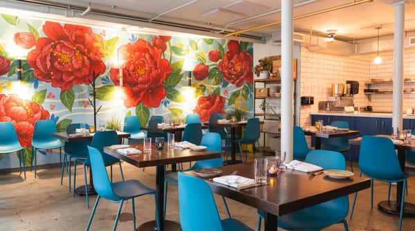 Restaurant dining room with a large red floral mural, dark wood tables, and bright blue chairs.