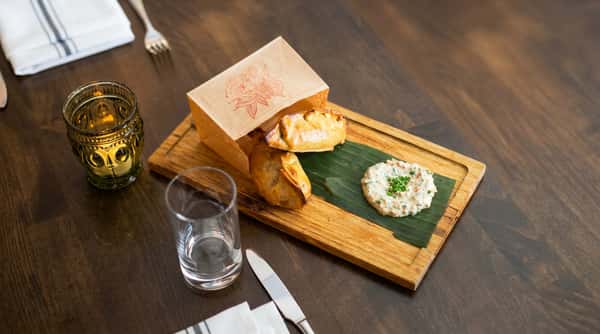 Two empanadas in a paper bag, a creamy dip on a banana leaf, and a glass of water on a wooden tray.