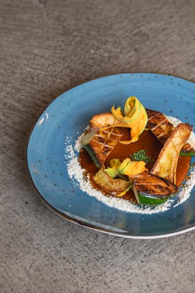 Grilled zucchini, yellow squash, and squash blossoms in a reddish sauce with white powder on a blue plate.