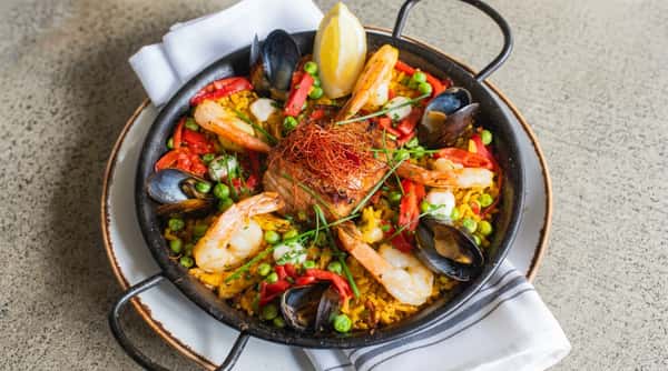Seafood paella with yellow rice, shrimp, mussels, red peppers, green peas, and a lemon wedge in a black pan.