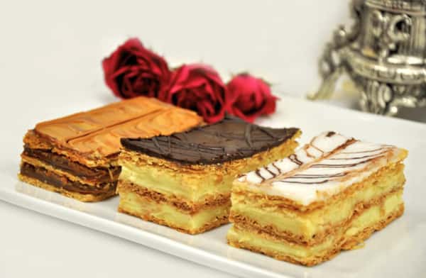 Assorted mille-feuille (Napoleon) pastries layered with flaky puff pastry and rich vanilla custard, topped with chocolate glaze and white icing drizzle, elegantly plated in a gourmet bakery setting.