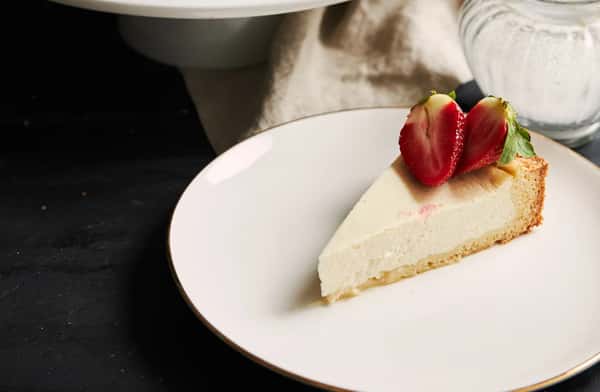 Slice of classic New York–style cheesecake topped with fresh strawberries on a white plate, highlighting a creamy texture and elegant café dessert presentation.