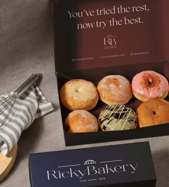 Open box of assorted gourmet donuts from Ricky Bakery, featuring powdered sugar, glazed, and chocolate-drizzled varieties, styled in branded packaging with elegant bakery branding for a premium dessert gift presentation.