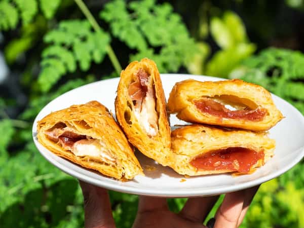 Flaky puff pastry turnovers filled with melted cheese and cured meat alongside a sweet fruit-filled pastry, served on a white plate outdoors, highlighting golden layers and a fresh café-style brunch presentation.