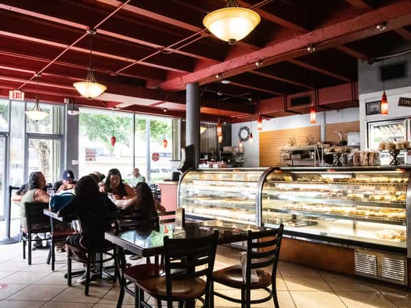 Cozy neighborhood café interior with customers dining at tables beside a glass pastry display case filled with fresh baked goods, warm pendant lighting, and large front windows creating an inviting bakery ambiance.