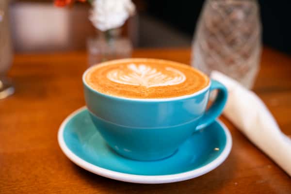A teal cup of latte with a leaf-shaped foam design sits on a matching saucer on a wooden table.