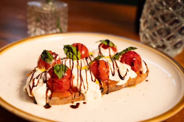 A slice of toast topped with roasted cherry tomatoes, fresh basil leaves, yogurt, and a drizzle of balsamic glaze.