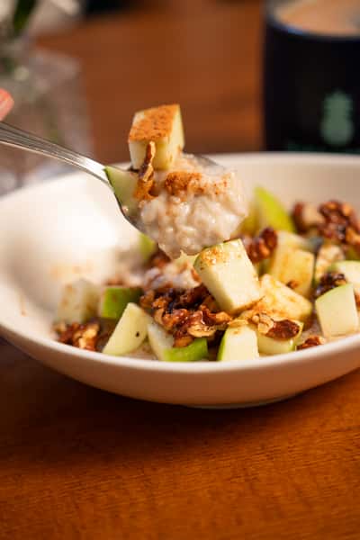 A spoon lifts a bite of oatmeal topped with green apple pieces, cinnamon, and granola from a bowl.