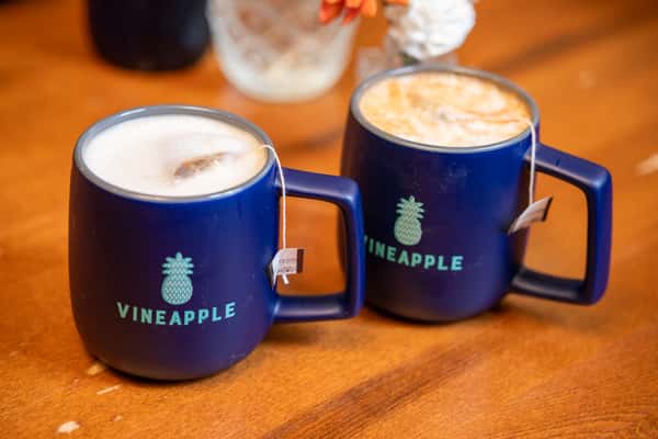 Two blue mugs with the word "VINEAPPLE" and pineapple logos, one containing hot tea and the other iced tea, on a wooden table.