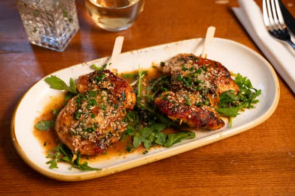 A close-up of a grilled chicken breast topped with chopped green onions, Parmesan cheese, and a drizzle of sauce, served on a bed of greens.