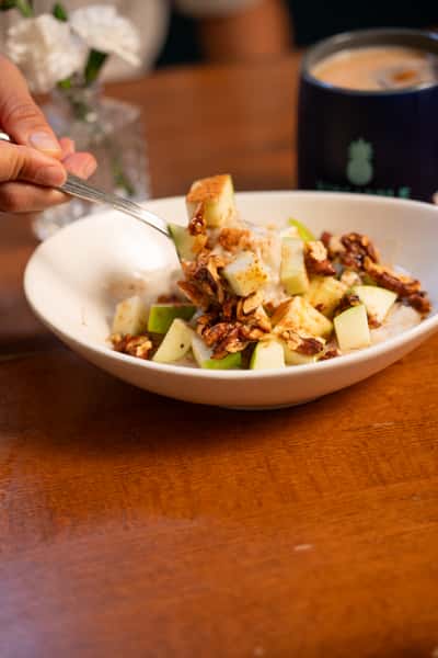 A spoon lifts a bite of oatmeal topped with green apple pieces, cinnamon, and granola from a bowl.