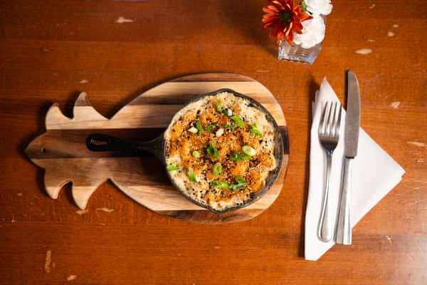 A cast iron skillet filled with baked macaroni and cheese, topped with breadcrumbs and green onions, served on a wooden board. A fork and knife are beside the dish.