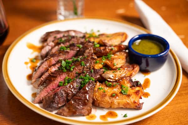 A plate of sliced steak garnished with chopped green onions, served with roasted potatoes and a small dish of green sauce.