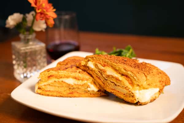 A grilled sandwich with layers of melted cheese, served with a side of fresh greens and a glass of wine in the background. The sandwich is cut in half, revealing the cheese inside.