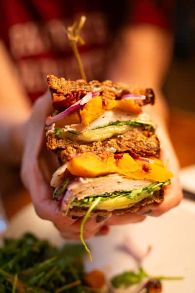 A hand holds a stacked sandwich with roasted turkey, grilled vegetables, arugula, red onion, and a slice of squash, secured with a toothpick.