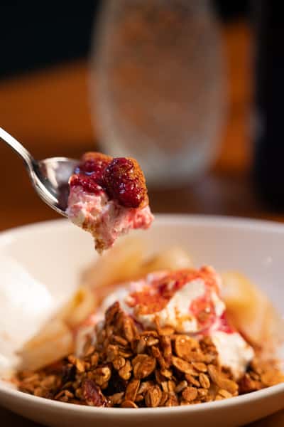 A spoon lifts a bite of creamy yogurt with raspberry sauce and granola from a bowl. Sliced pears are visible in the background.