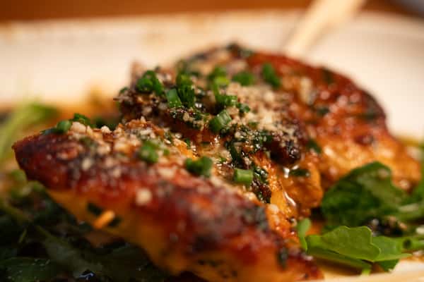 A close-up of a grilled chicken breast topped with chopped green onions, Parmesan cheese, and a drizzle of sauce, served on a bed of greens.