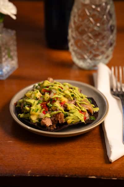 A black corn tortilla topped with grilled meat, fresh salsa, avocado sauce, and cilantro.