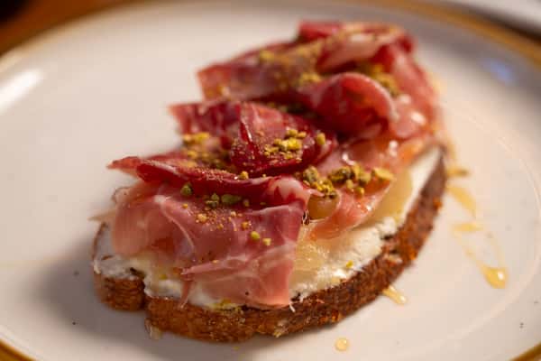 A slice of toasted bread topped with cream cheese, prosciutto, crushed pistachios, and a drizzle of honey.