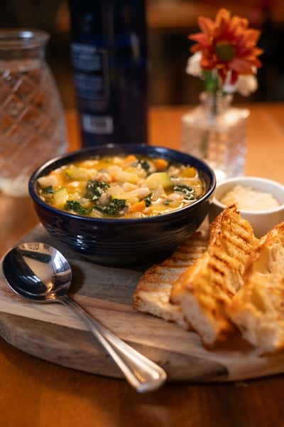 A bowl of vegetable soup with white beans, kale, and carrots, served with grilled bread and a small dish of butter.