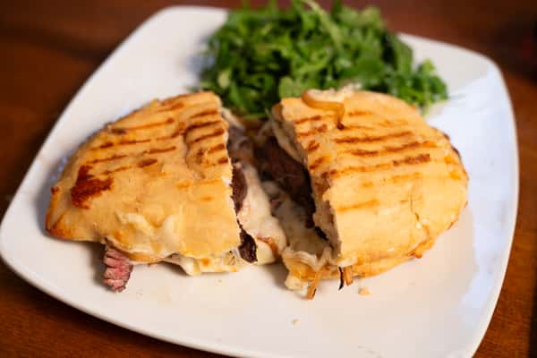 A grilled sandwich filled with roast beef and melted cheese, served with a side of arugula salad. The sandwich is cut in half, revealing the filling.