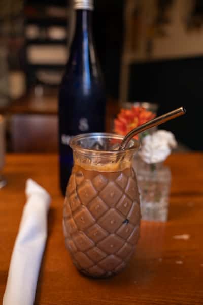 A glass shaped like a pineapple holds iced chocolate with a metal straw. A bottle and flowers are blurred in the background.