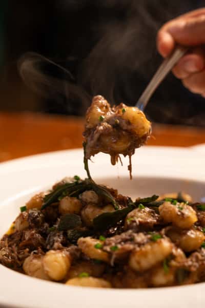 A person lifts a spoonful of gnocchi with slow-cooked beef, sage, and grated cheese from a plate. Steam rises from the dish.