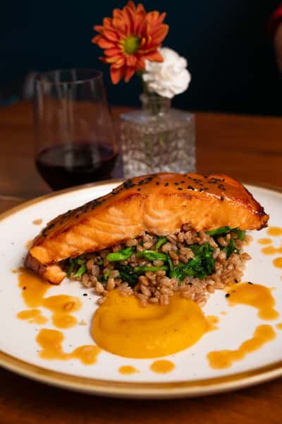 A salmon fillet sits atop farro and sautéed greens, accompanied by a swirl of butternut squash puree and drizzled with sauce. A glass of red wine is blurred in the background.