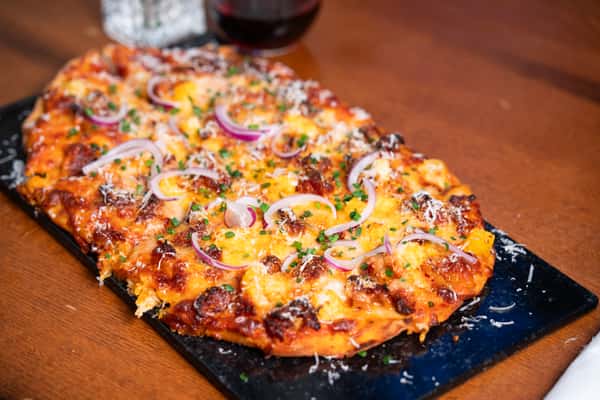 A pizza topped with melted cheese, sausage, red onion slices, chopped chives, and grated Parmesan, served on a dark stone plate.