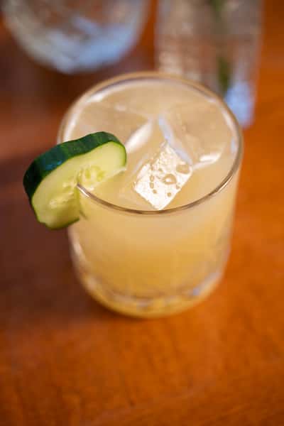 A refreshing drink with ice cubes and a cucumber slice garnish, served in a short glass.