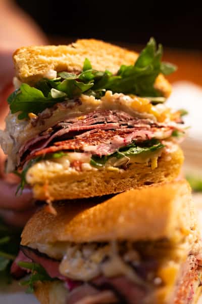 A hand holds a stacked sandwich with layers of pastrami, leafy greens, and creamy dressing between slices of toasted bread.