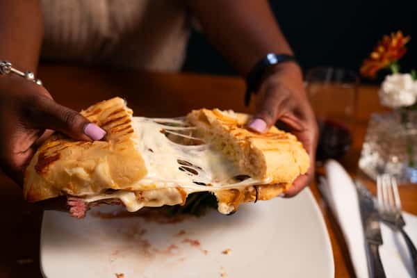 A person pulls apart a grilled sandwich filled with melted cheese, revealing the gooey interior, with a side of salad and a glass of wine in the background.