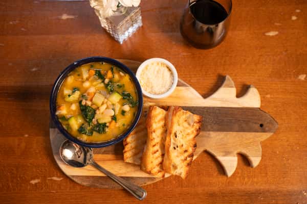 A bowl of vegetable and white bean soup with spinach, served with grilled bread and a side of grated Parmesan cheese on a wooden serving board.