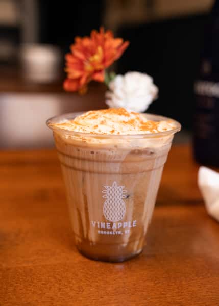 An iced coffee topped with whipped cream and a sprinkle of cinnamon, served in a clear cup with a "Vineapple" logo. A flower is in the background.