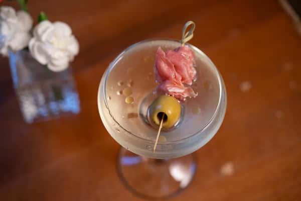 A classic martini garnished with an olive and a slice of prosciutto on a cocktail pick, served in a chilled glass.