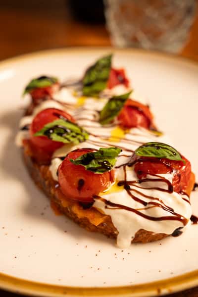 A slice of toast topped with roasted cherry tomatoes, fresh basil leaves, yogurt, and a drizzle of balsamic glaze.