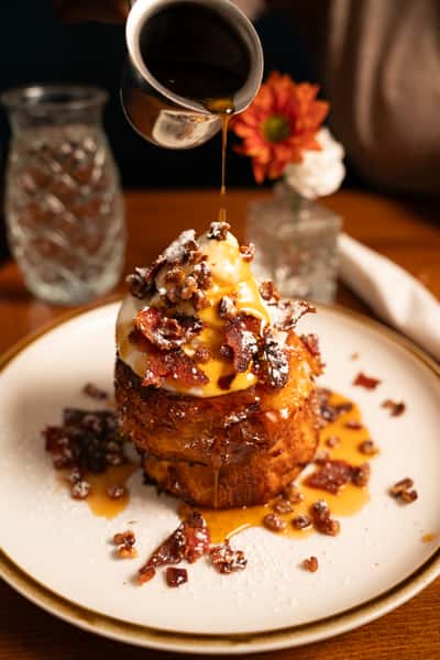 Thick French toast topped with whipped cream, candied pecans, crispy bacon, and maple syrup being poured over.