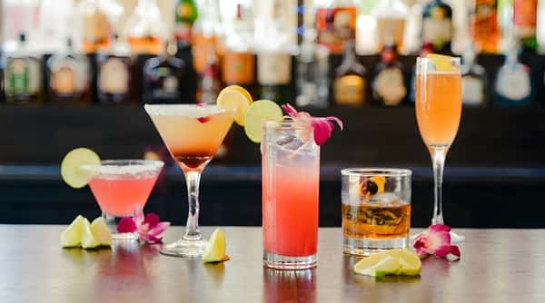 Five colorful cocktails and lime/lemon wedges on a smoky bar counter with blurred liquor bottles in background.