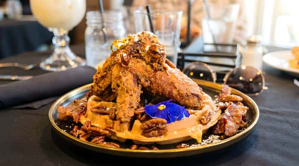 Fried chicken and waffle dish on a black plate with pecans, powdered sugar, and a blue edible flower.