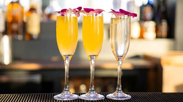 Three champagne flutes with yellow and clear cocktails, garnished with purple orchids, on a dark bar with blue lighting.