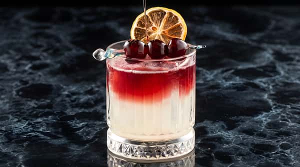 Layered cocktail in a cut glass, with a red top and white bottom, garnished with cherries and a dried lemon slice.