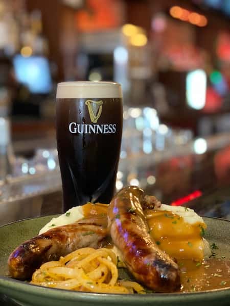A pint of cold Guinness sits on the bartop next to a hot plate of Irish Bangers and Mashed Potatoes 