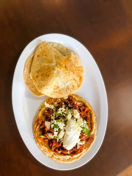 Traditional Mexican sope topped with seasoned chorizo, crumbled queso fresco, diced onions, cilantro, and sour cream, served with warm handmade gorditas on a white plate.
