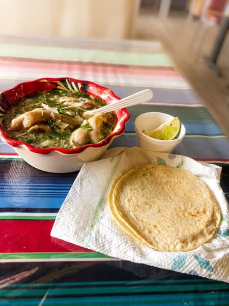 Traditional Mexican chicken soup with vegetables, hominy, fresh herbs, lime wedges, and warm corn tortillas on a colorful tablecloth.